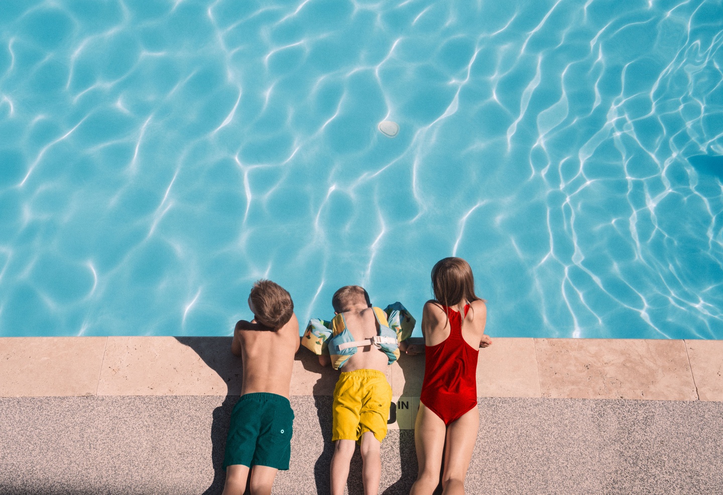Three children in swimwear lie at pool's edge, with vibrant blue water.