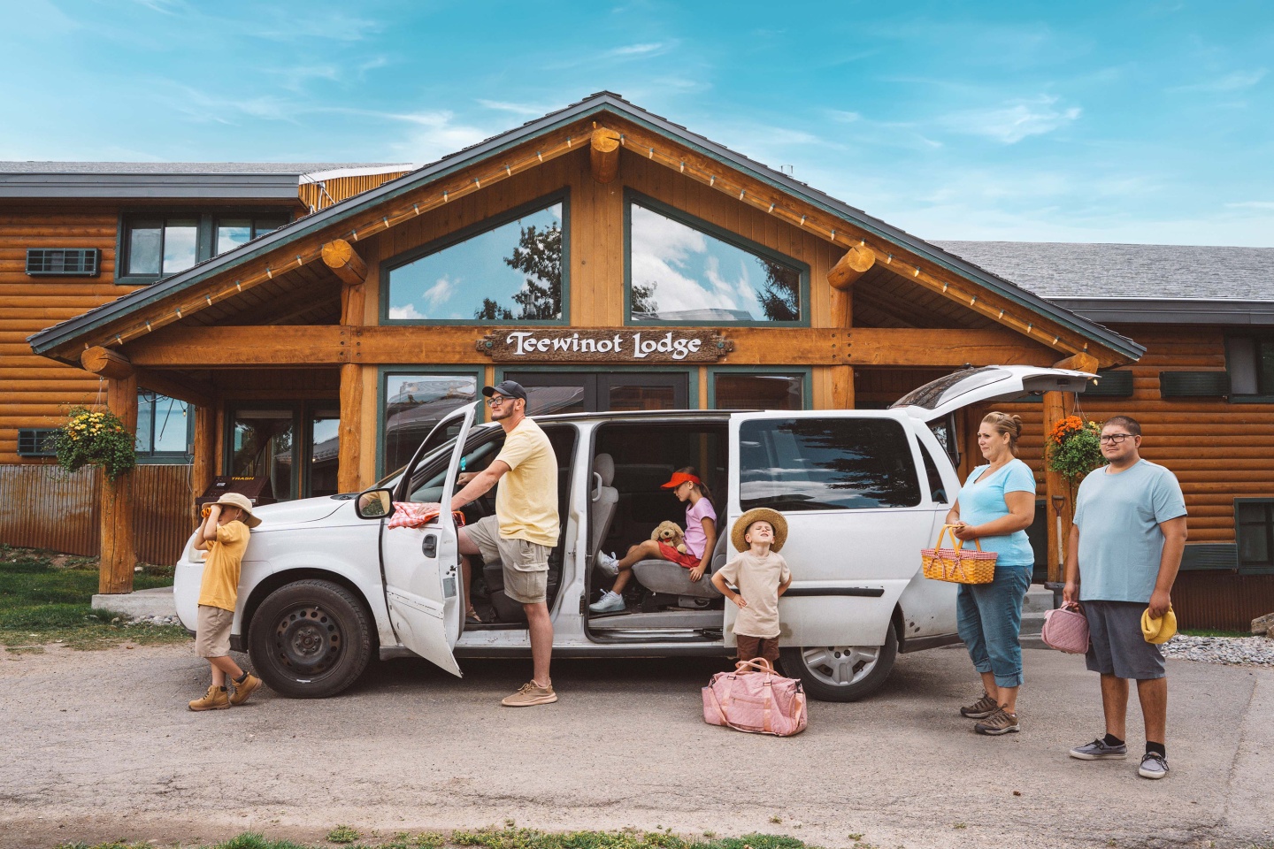 Family with van packed at a log cabin lodge, children holding colorful bags.