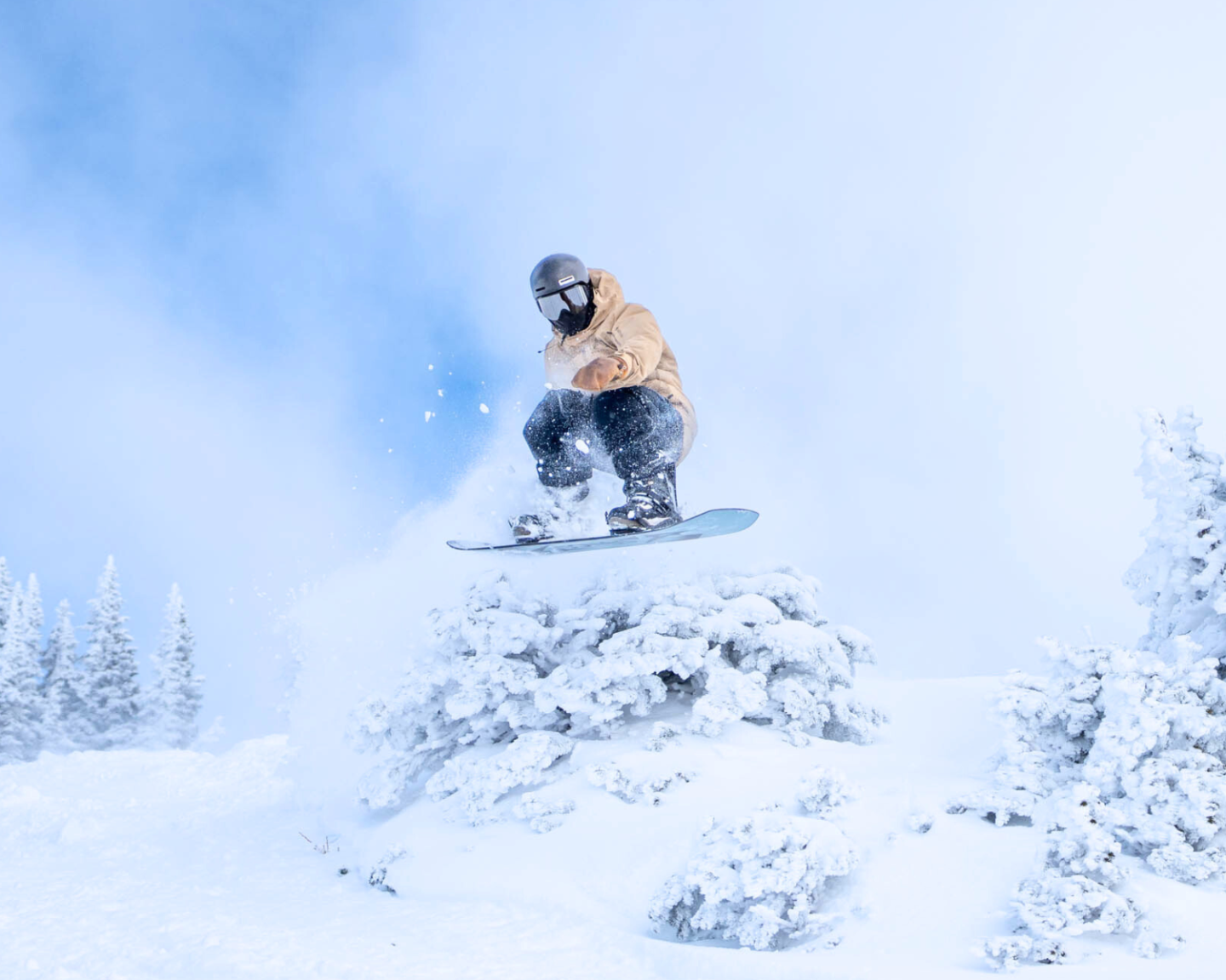 Snowboarder in mid-air jump above snowy trees, wearing a helmet and beige jacket.