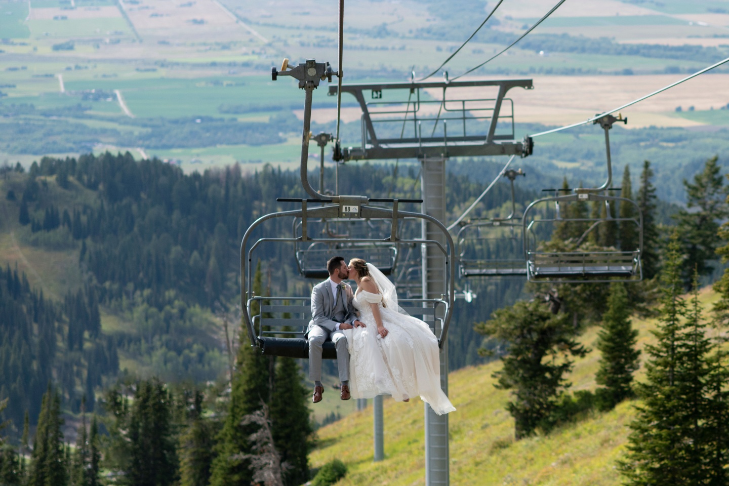 Bride and groom kiss on a ski lift above a forested mountainside.