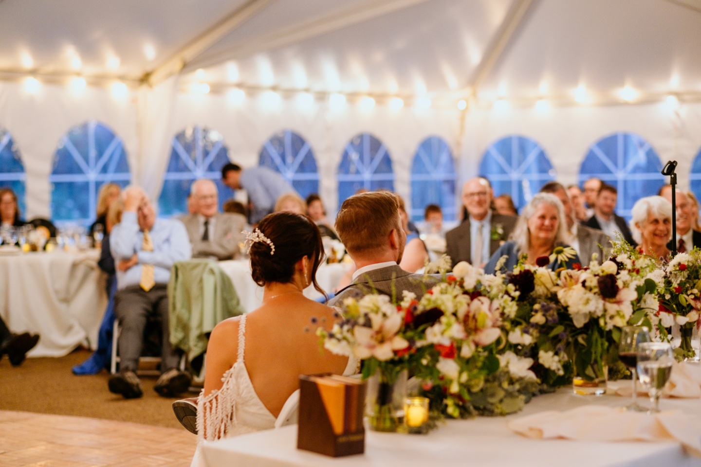 Wedding reception with guests seated at decorated round tables under a white tent.