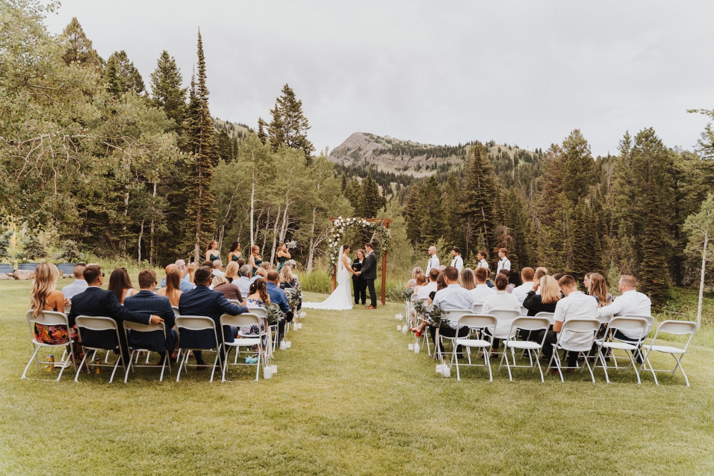 Outdoor wedding ceremony surrounded by trees and mountains, guests seated on folding chairs.