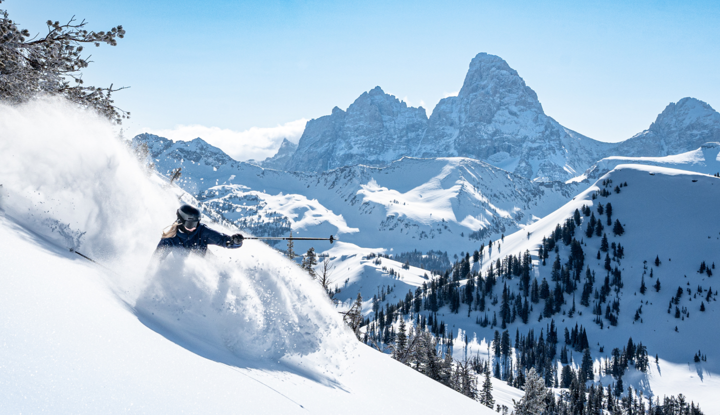 Skier navigating powder on a snowy mountain slope, clear sky, distant peaks.