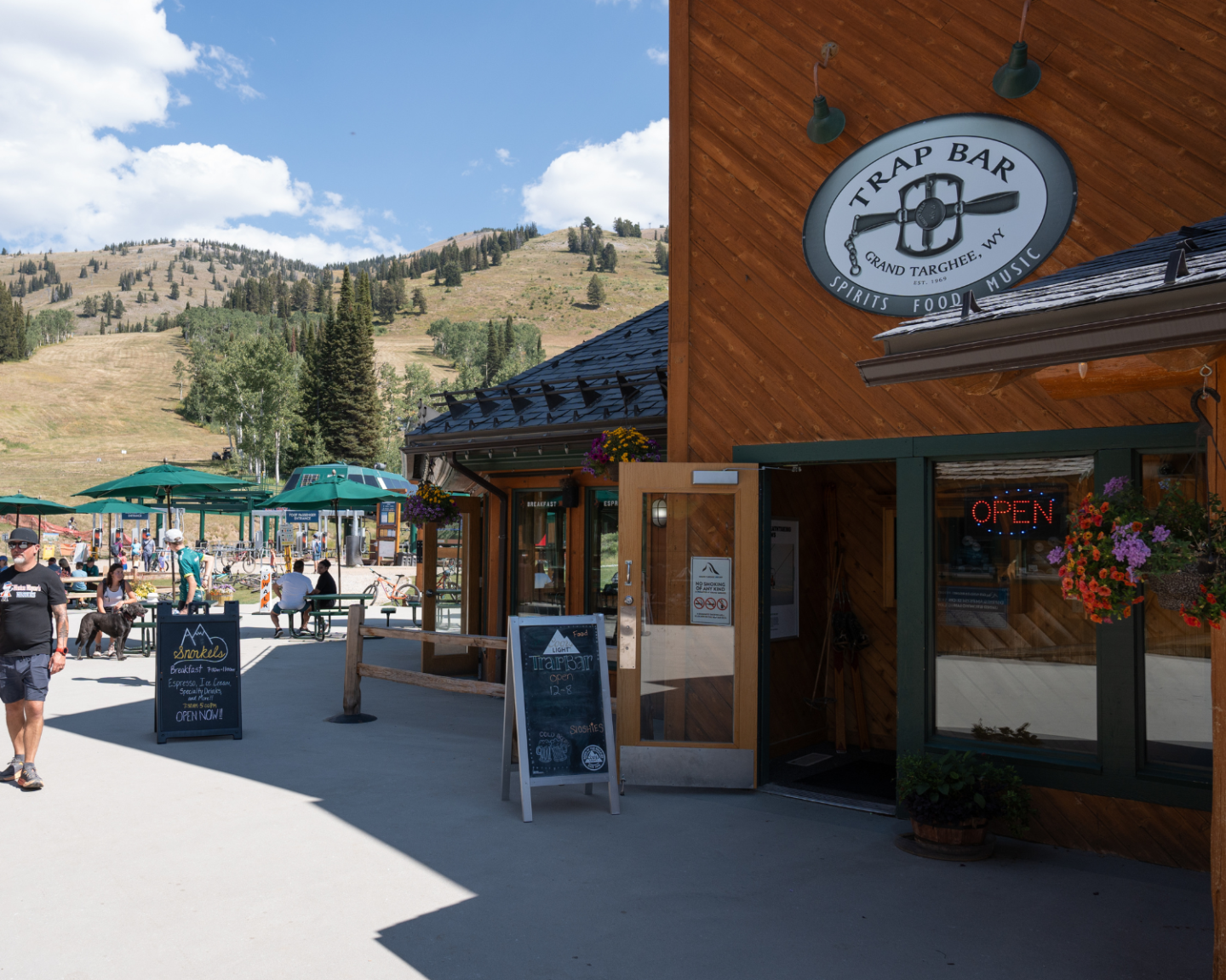 Mountain bar entrance with outdoor seating, sunny day, and people walking.