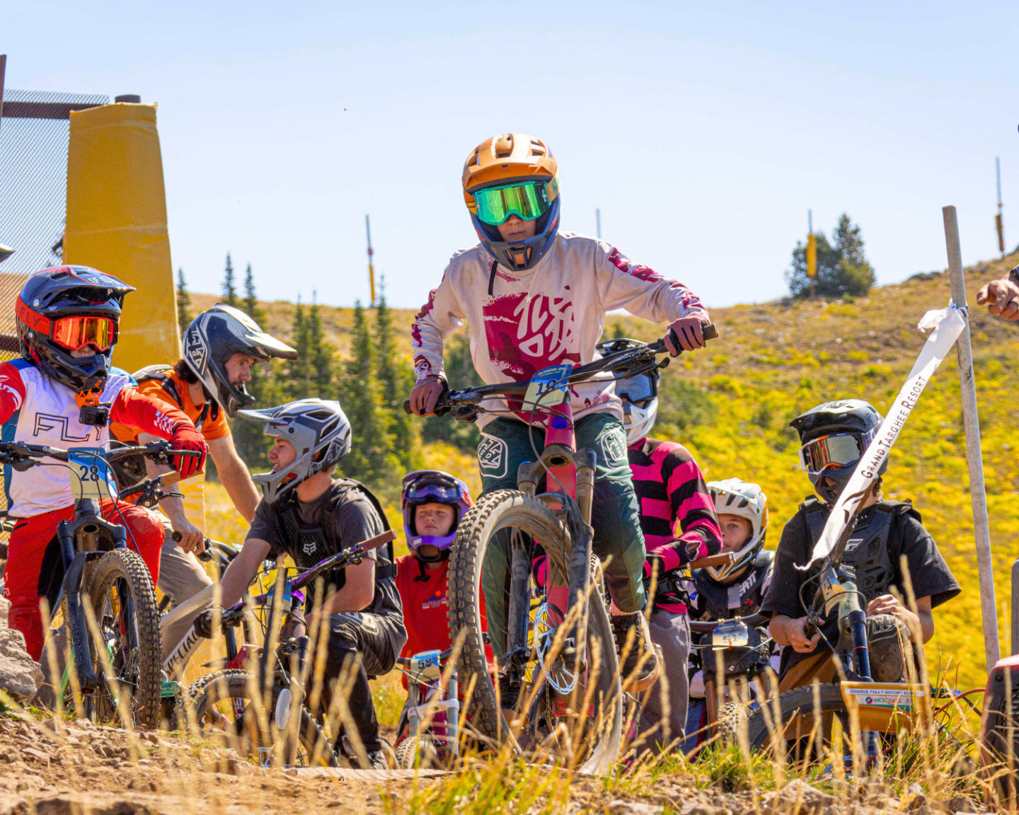 Children on mountain bikes at starting line, wearing helmets and colorful gear.