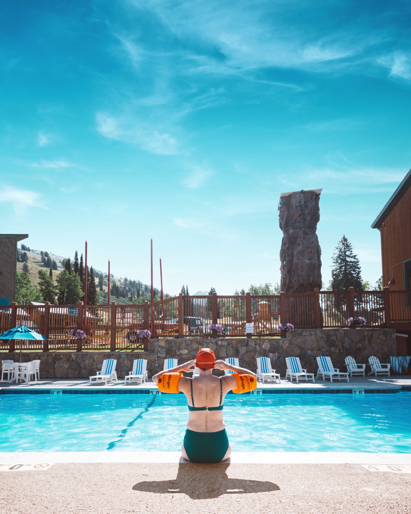 Person in pool, arm floats on, under bright blue sky, surrounded by buildings and lounge chairs.