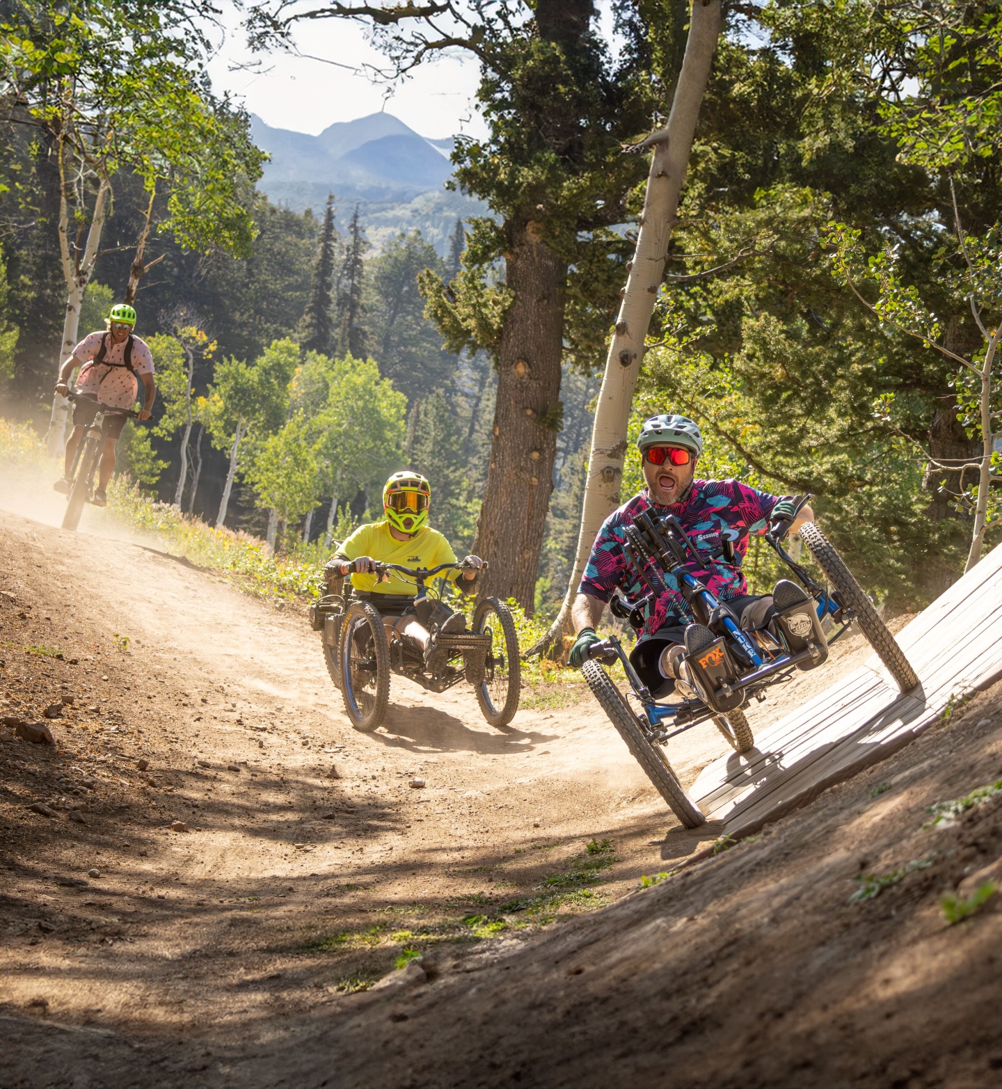 Three people riding adaptive bikes on a dirt trail in a forest.