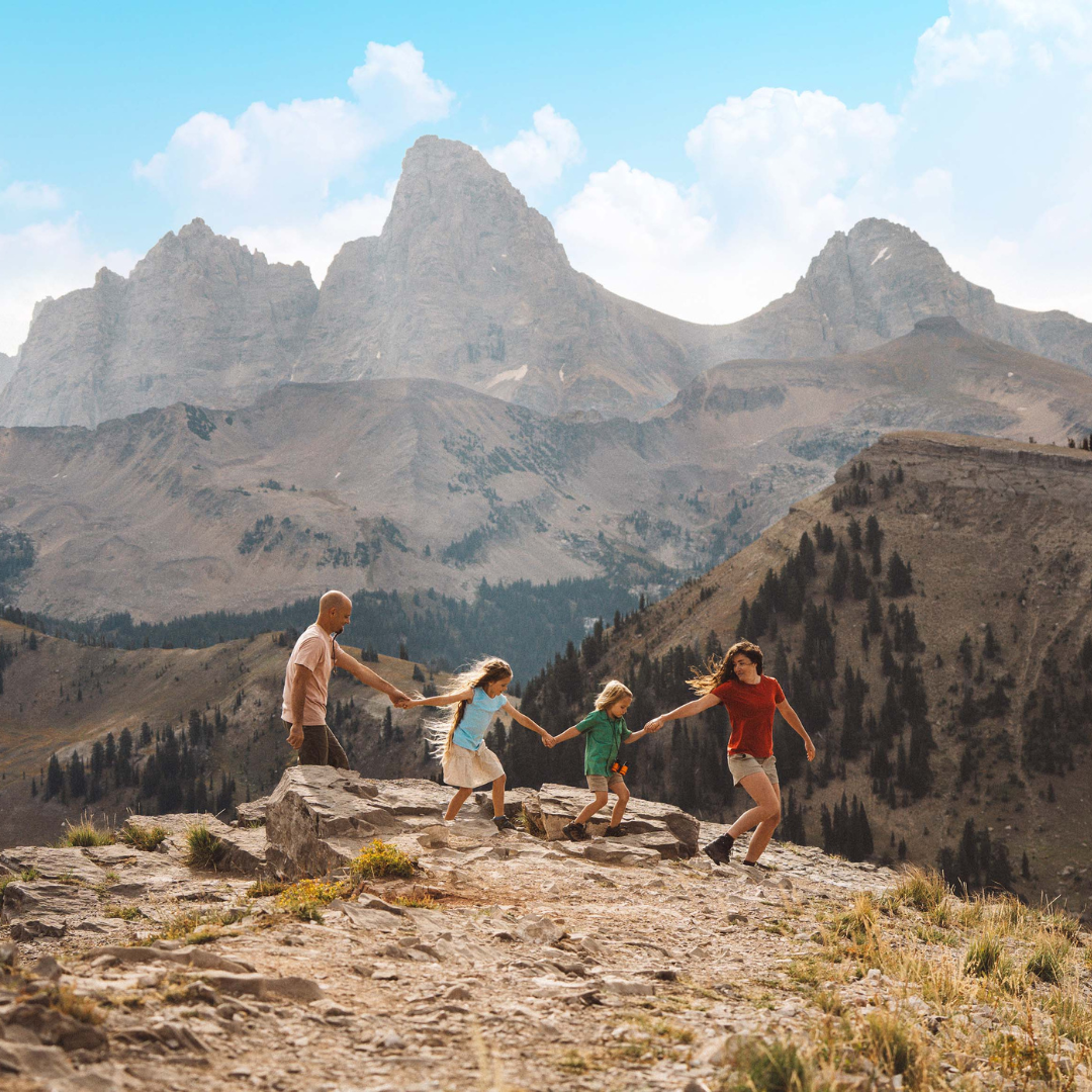 Family holding hands, hiking on a rocky mountain path against a backdrop of tall peaks.