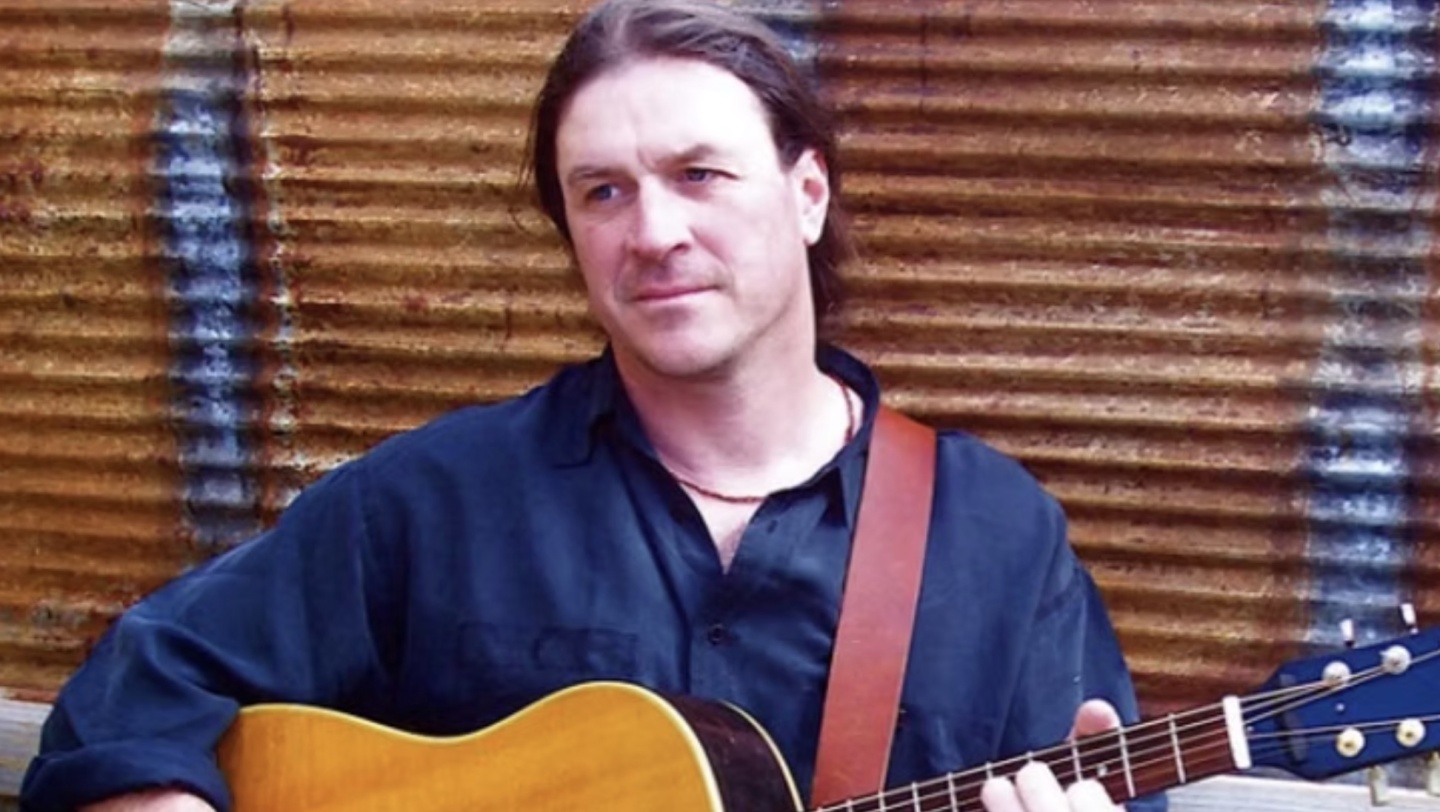 Man in a dark shirt playing acoustic guitar, corrugated metal background.