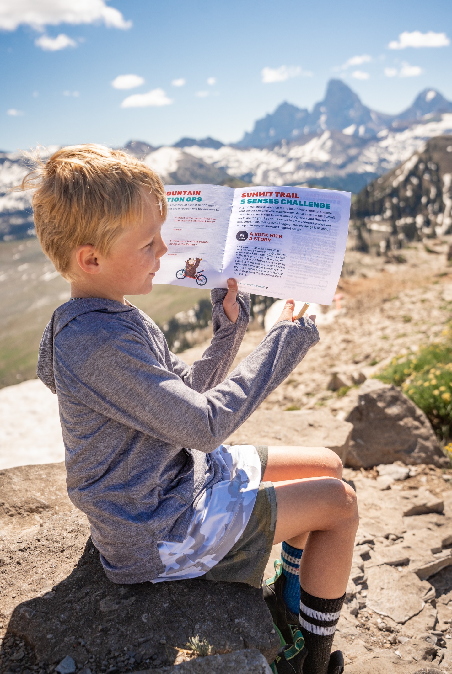 Child reading paper on rocky hill with mountains in background.