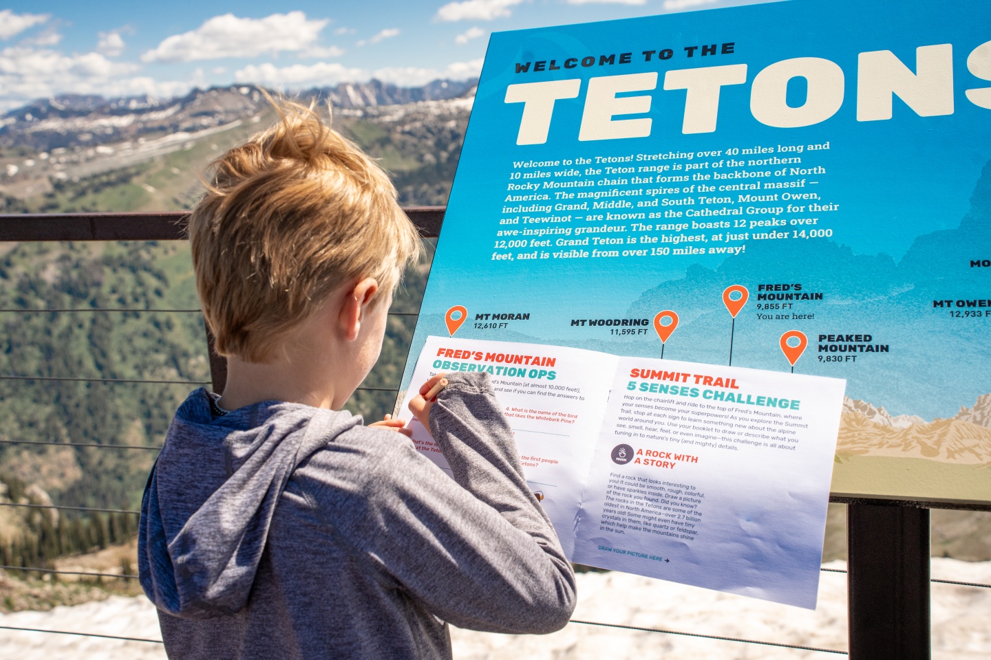 A child in a hoodie reads an informational sign at a mountain viewpoint.