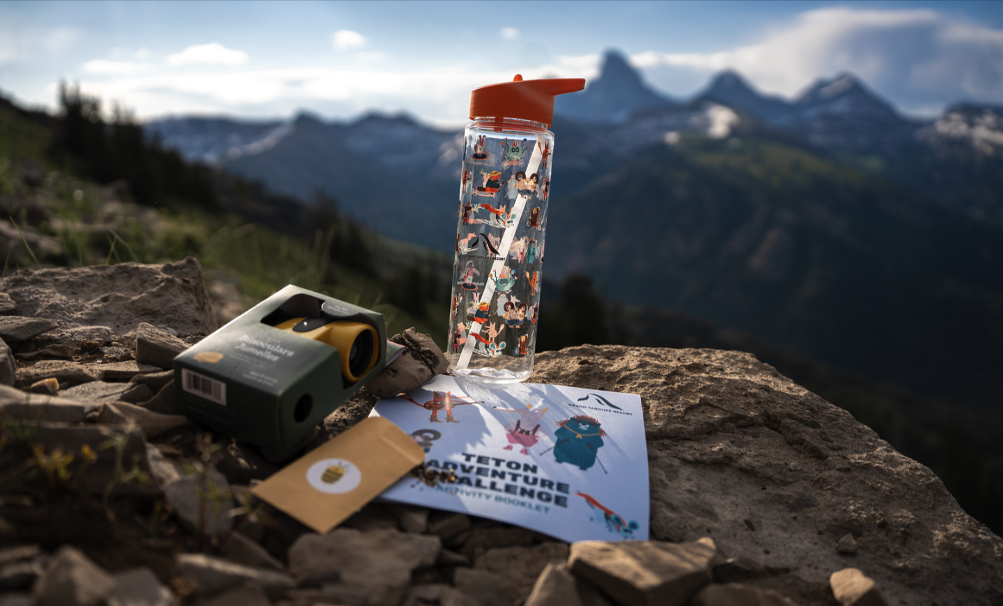 Camping gear with a water bottle and map on a rocky mountain overlooking peaks.