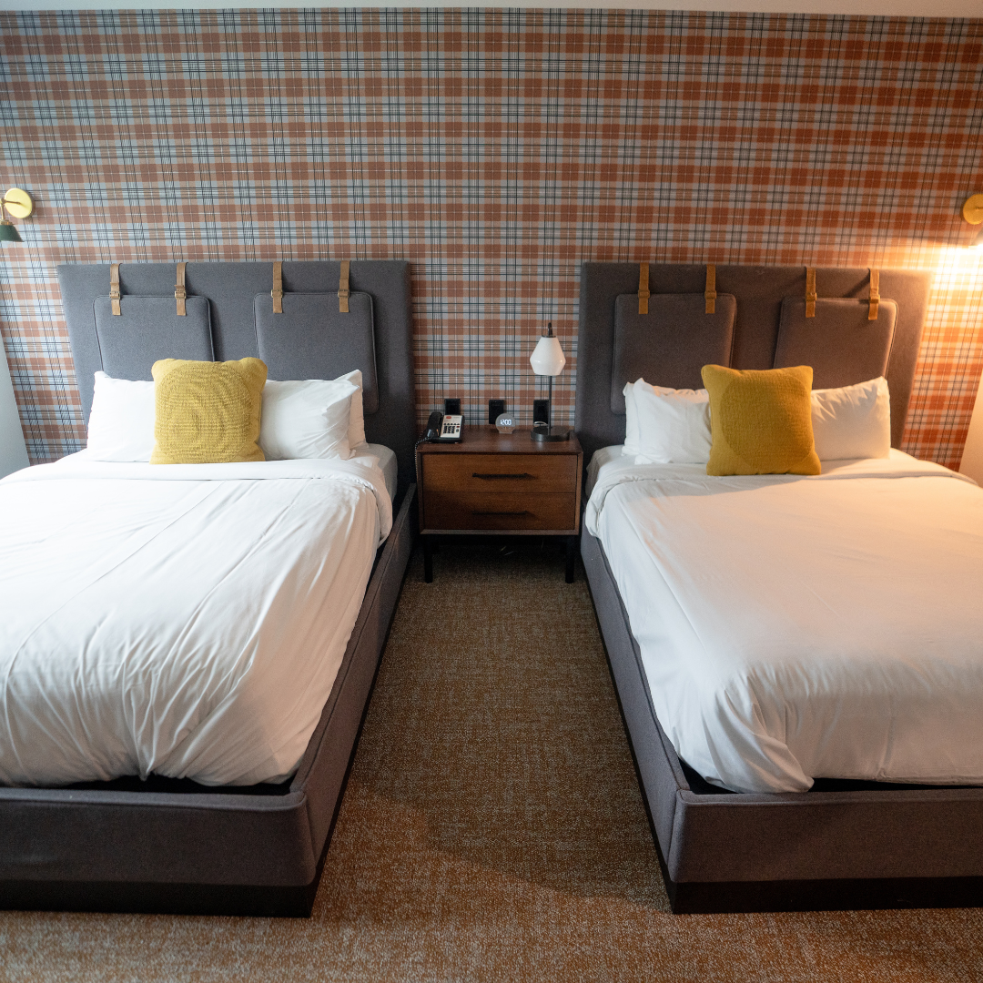 Two neatly made beds with yellow cushions in a hotel room with plaid wallpaper.