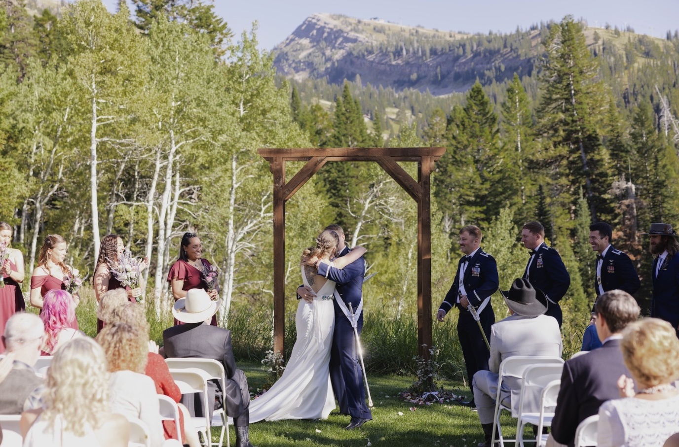 Wedding ceremony outdoors with a couple embracing, surrounded by guests and mountains in the background.
