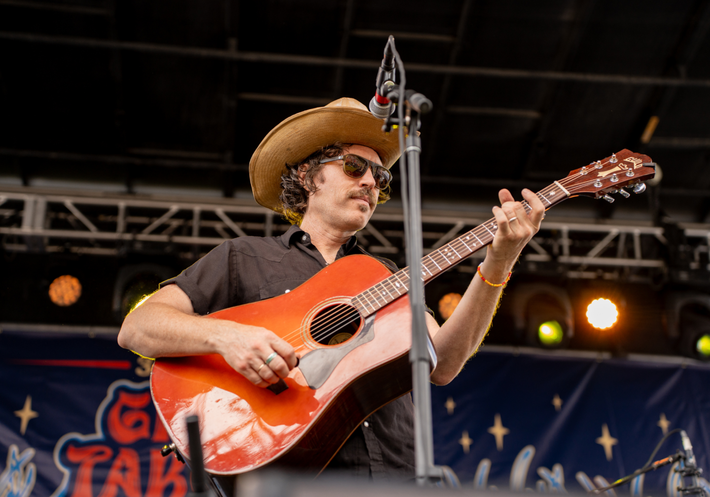 Musician with guitar on stage, wearing sunglasses and a cowboy hat.