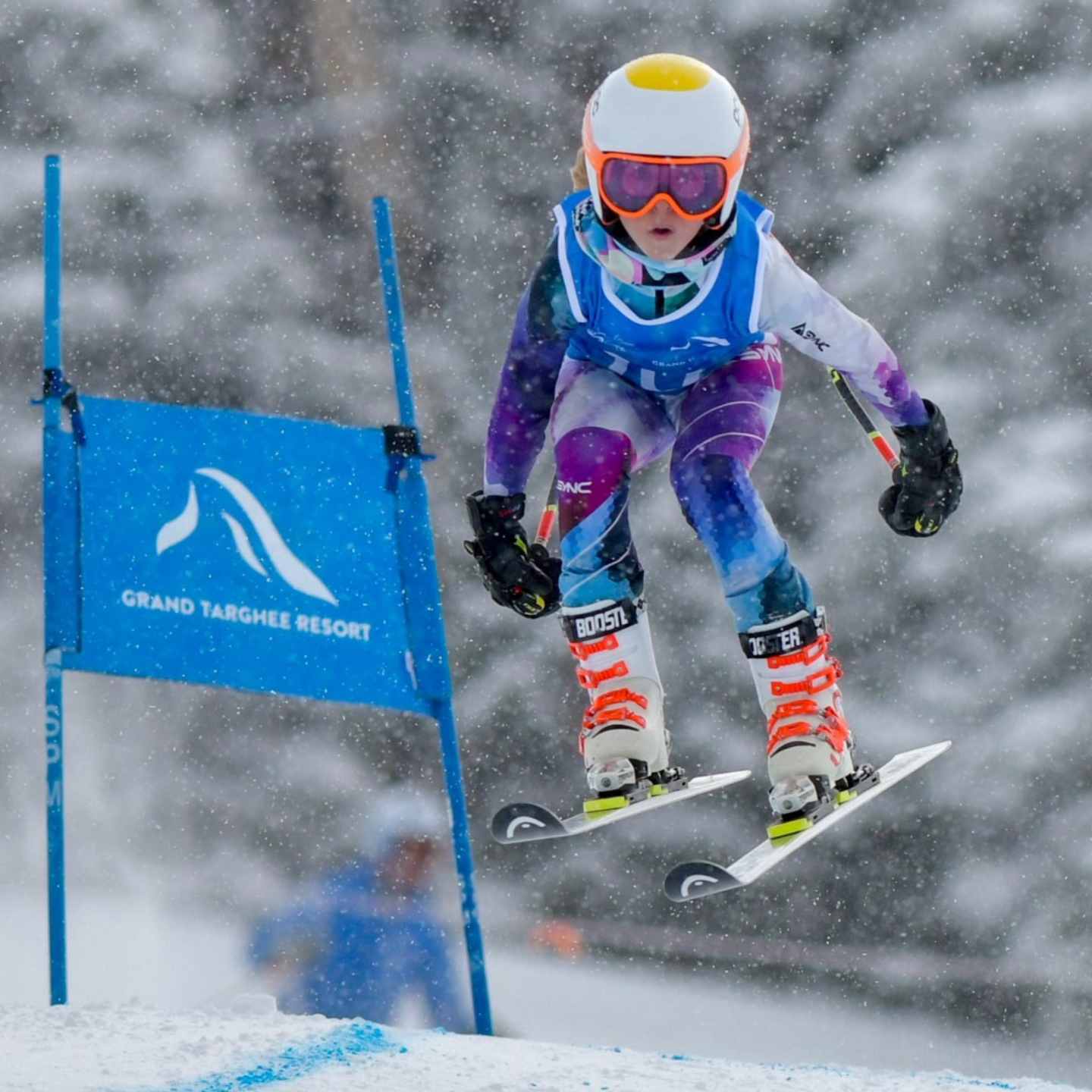 Youth skier jumping mid-air, wearing colorful gear and helmet, snowy background.