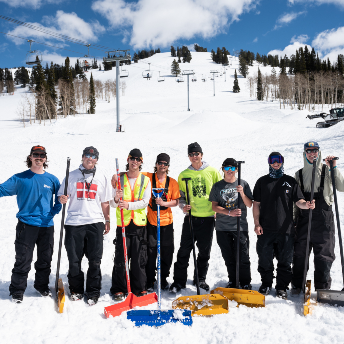Seven people with shovels on a snowy hill under a blue sky.
