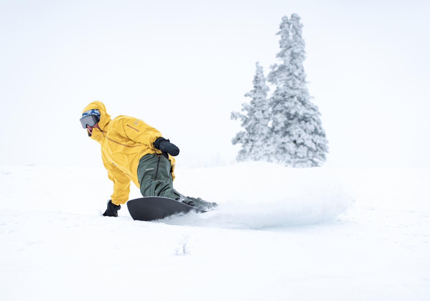 Snowboarder in a yellow jacket carving through fresh snow, with snowy trees in the background.