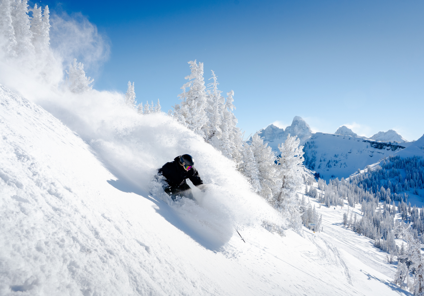 Skier carving through powder on a snowy mountain slope, clear blue sky.