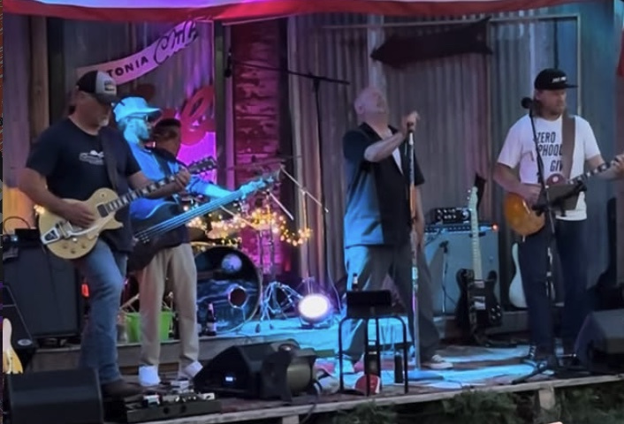 Band performing on stage with colorful lights and rustic backdrop.