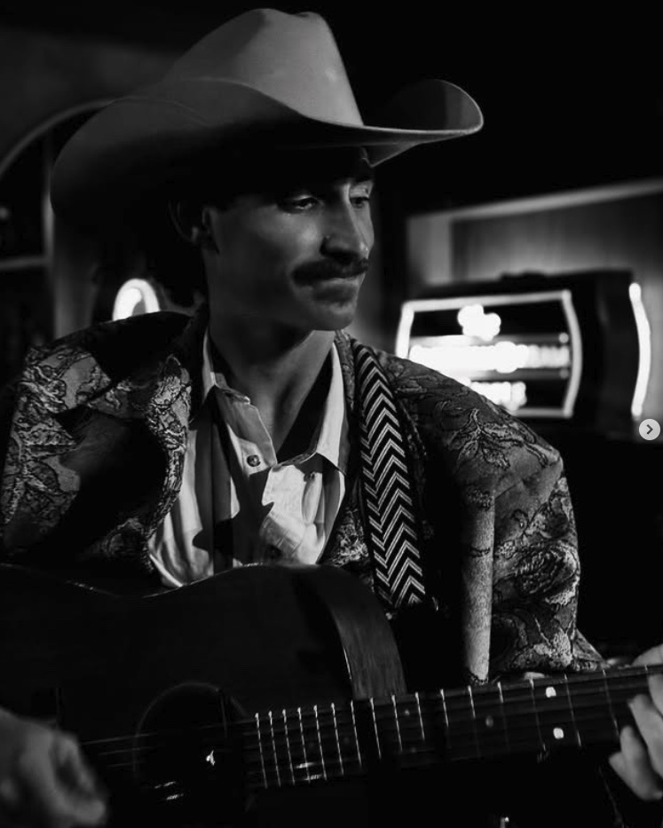 Cowboy playing guitar in dimly lit bar, wearing a patterned jacket and hat. Black and white photo.