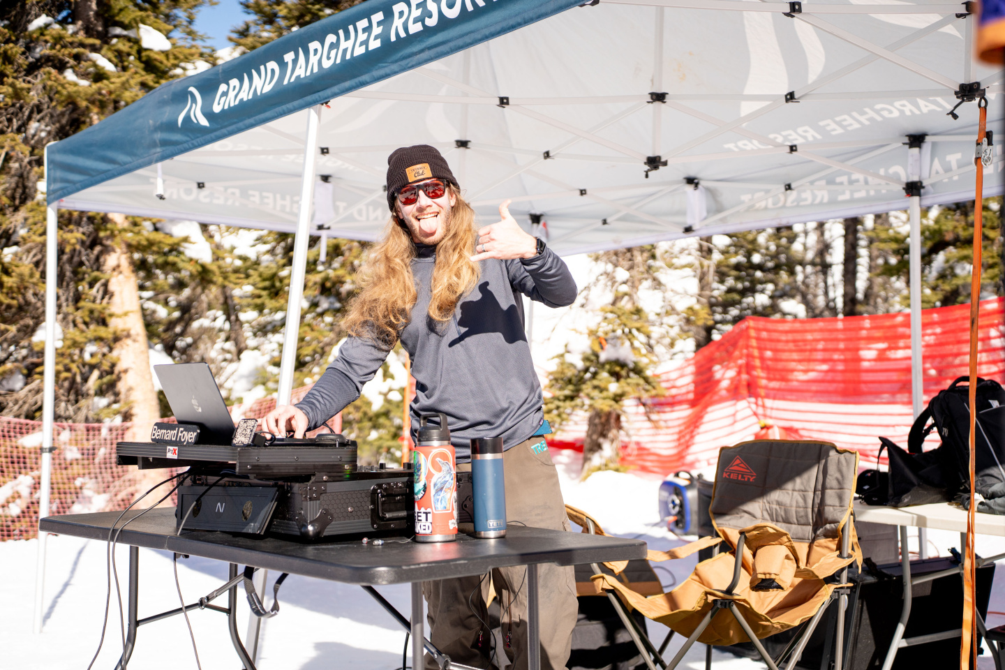 DJ under a tent mixes music on a snowy day, wearing headphones and sunglasses.