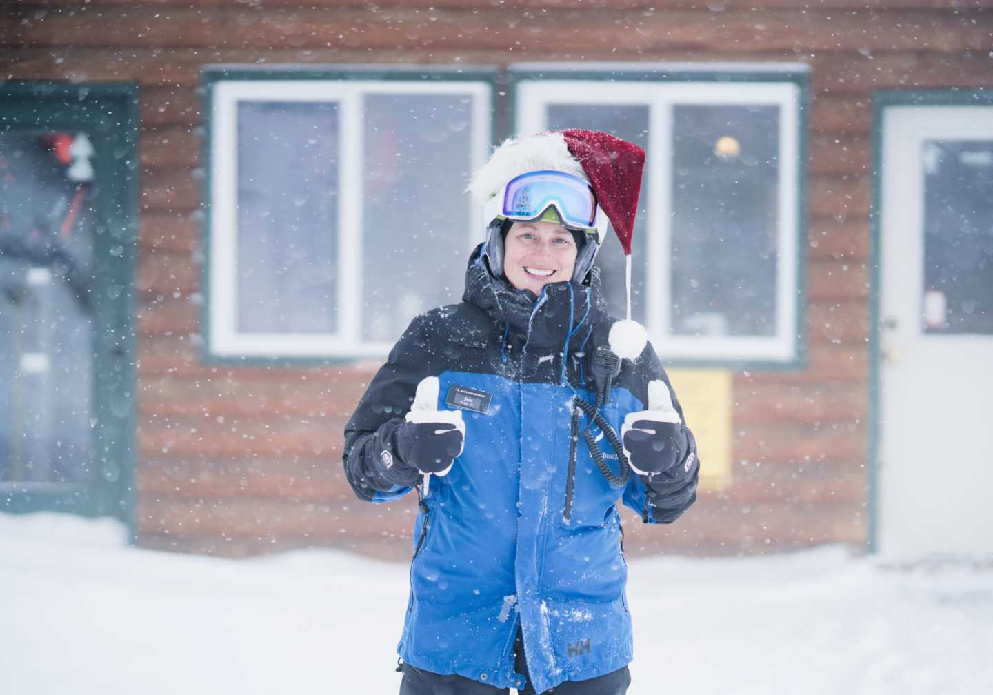 Person in snow gear giving thumbs up, wearing a Santa hat, snowy background.