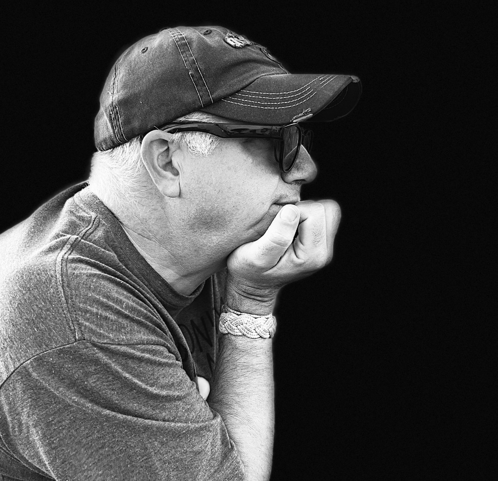 Man in profile wearing cap and sunglasses, hand on chin, against black background.