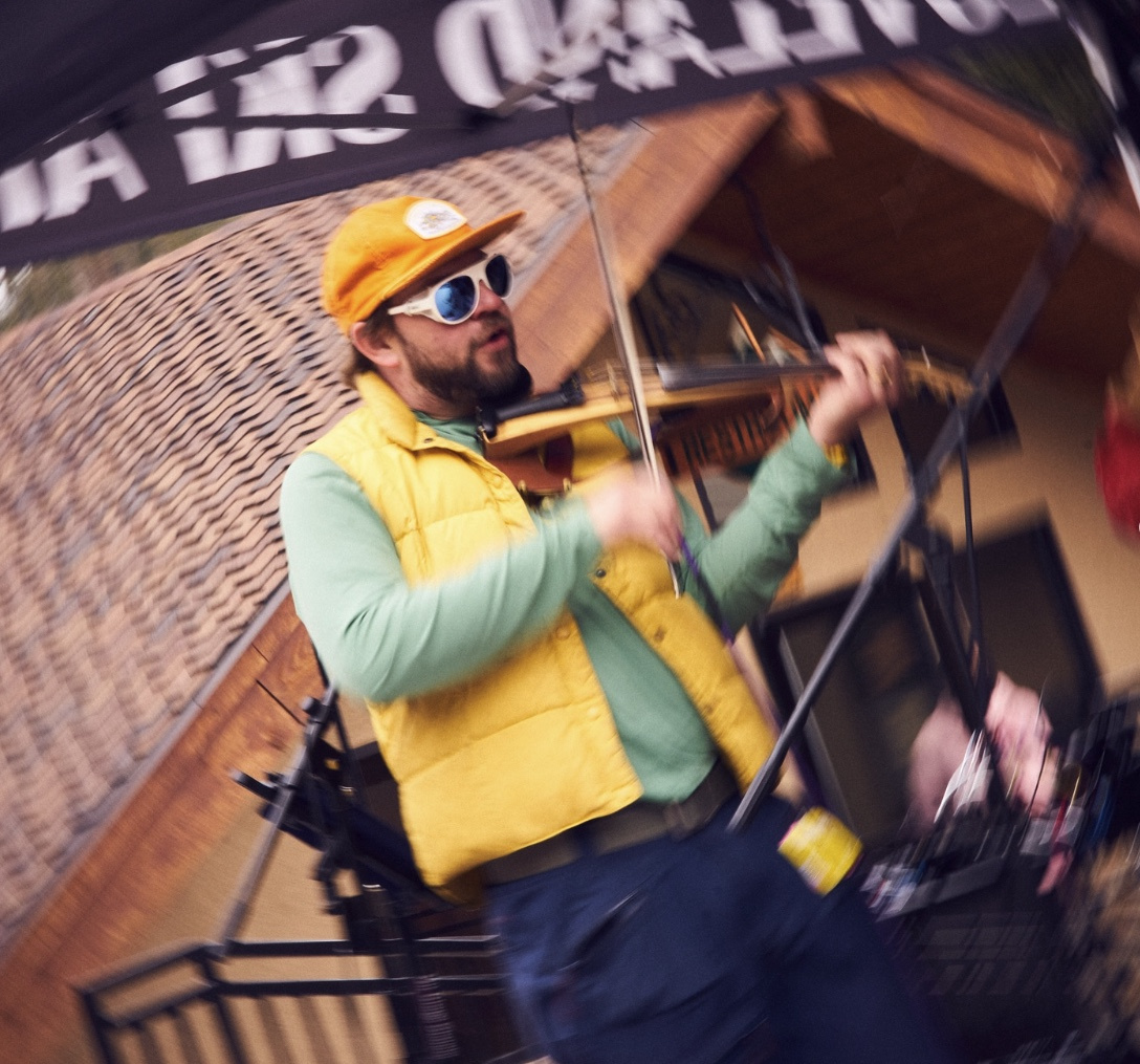 Man in yellow vest and cap playing violin outdoors.