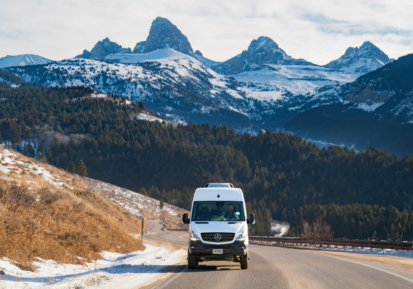 A van drives on a scenic mountain road with snow-covered peaks in the background.