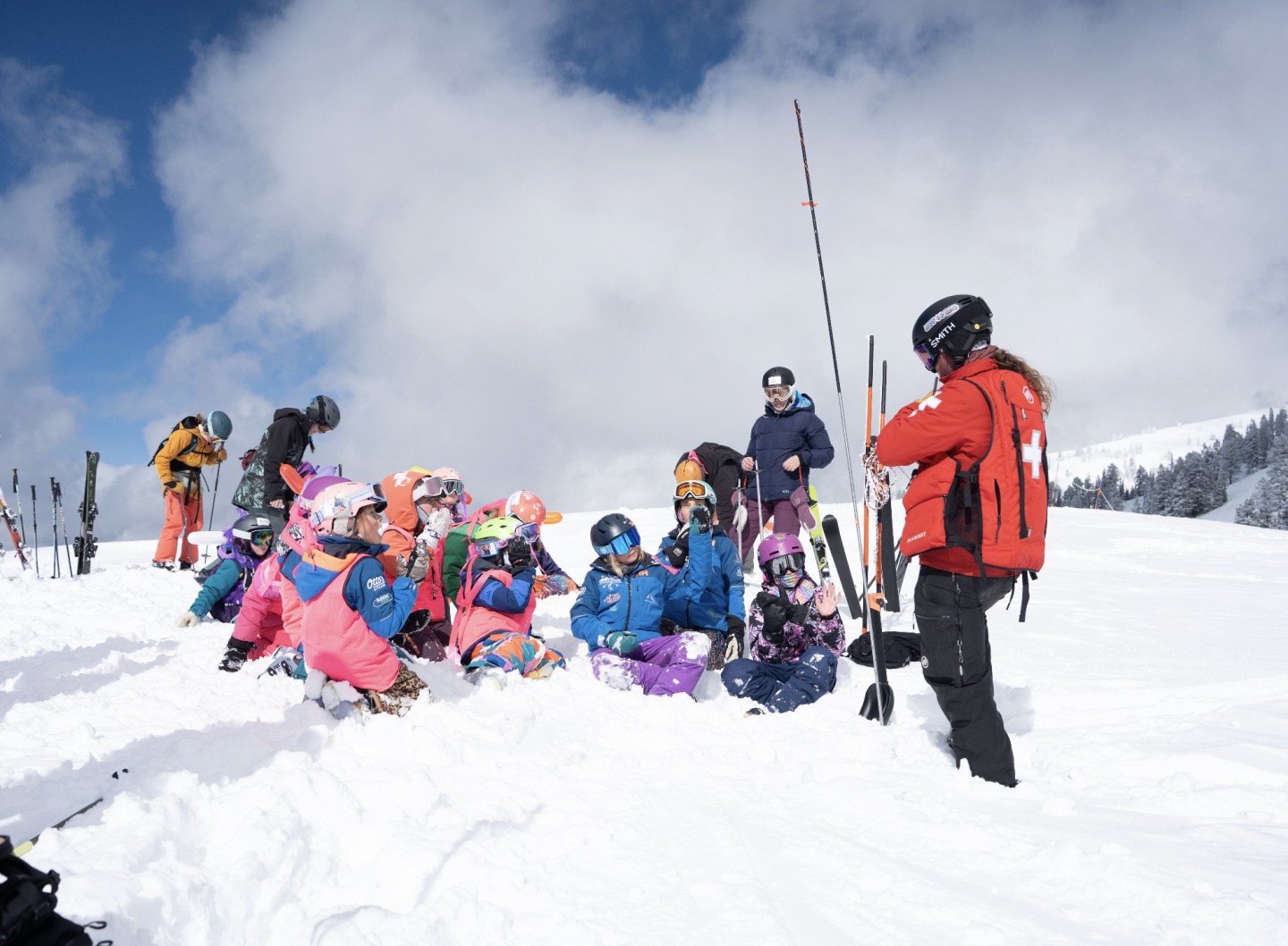 Children in colorful snowsuits gather around a ski instructor on a snowy mountain slope.