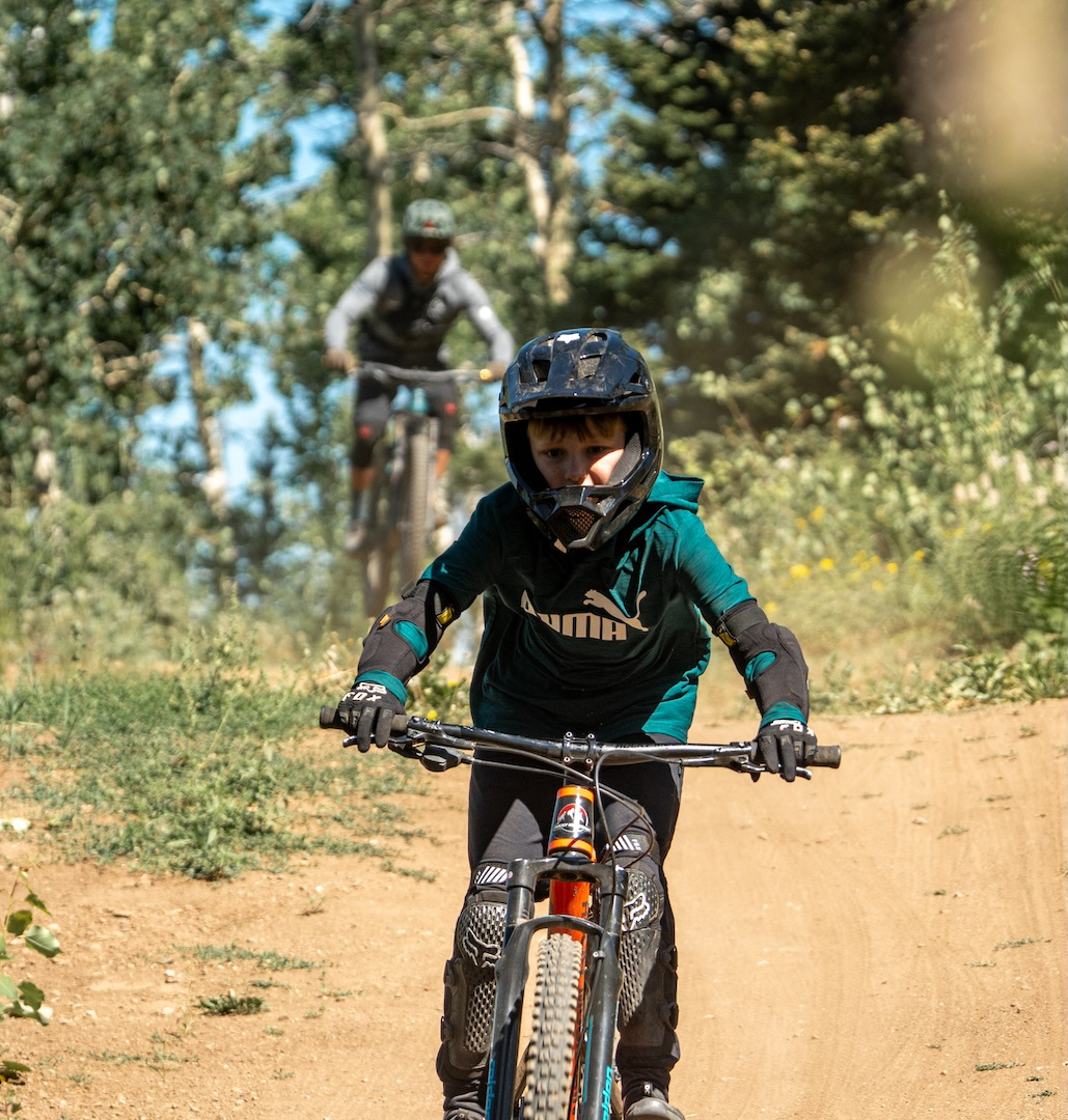 Mountain bikers on a forest trail, one in focus wearing a helmet and teal jersey.