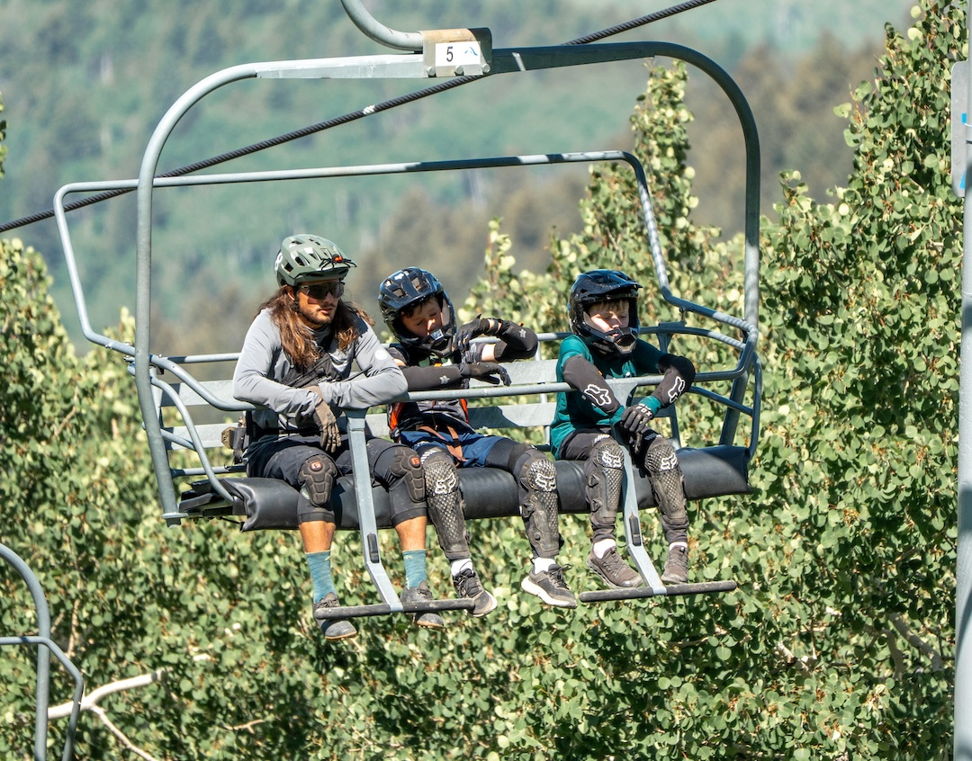 Three people in helmets on a ski lift, surrounded by green trees.