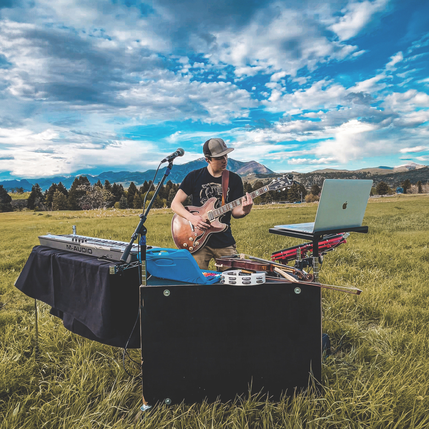 Musician playing guitar in an open field with equipment and mountains in the background.