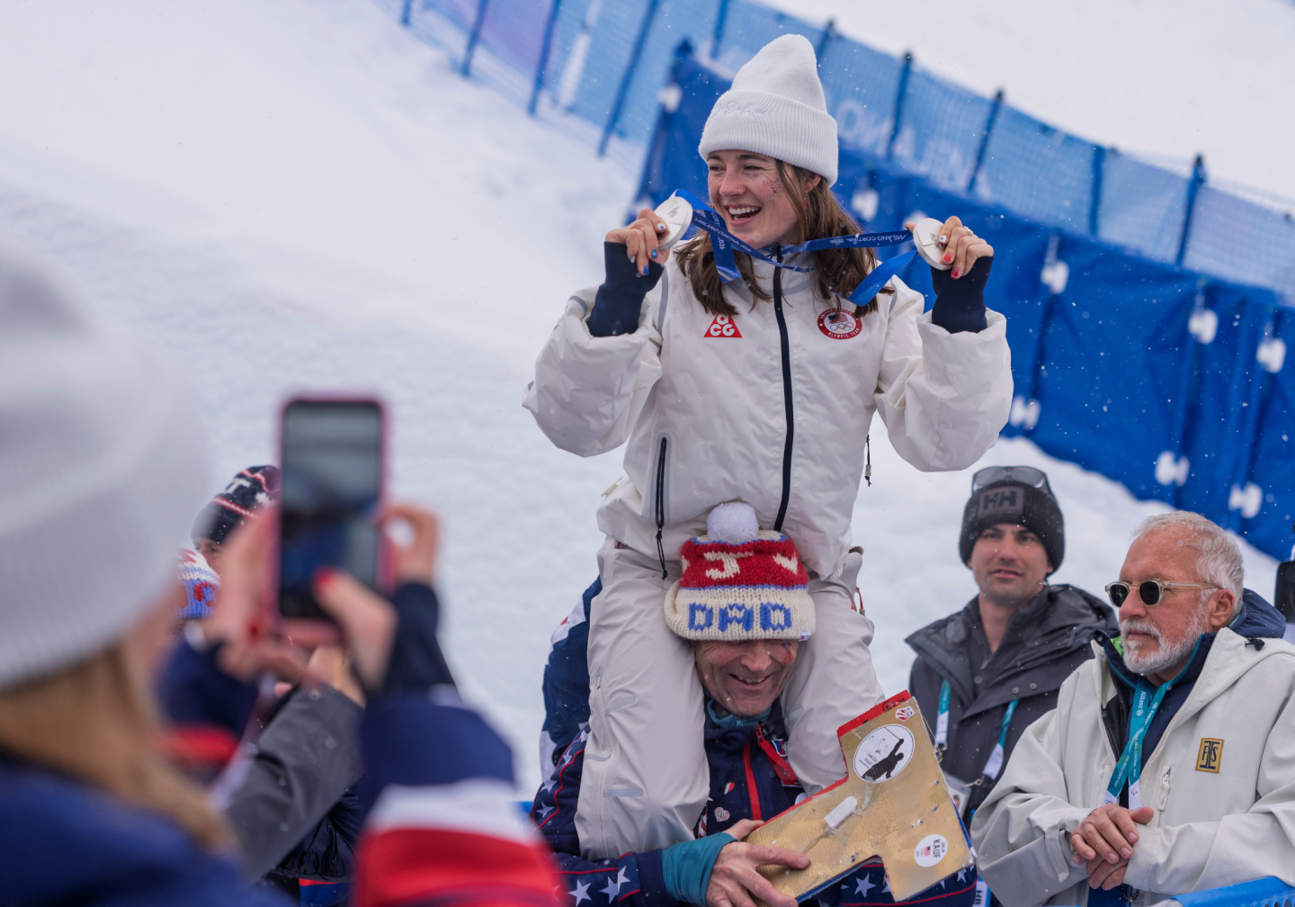 Young athlete in winter attire, joyfully holding medals on snowy shoulders of a supporter.