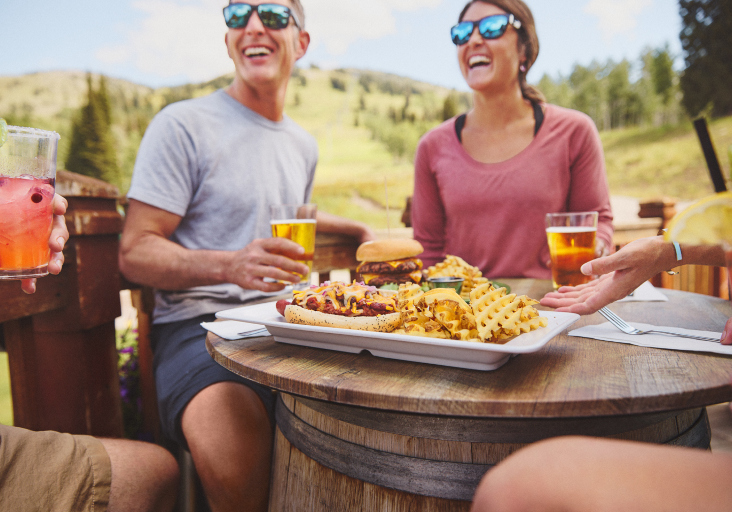 Friends enjoying food and drinks outside on a sunny day, laughing together.