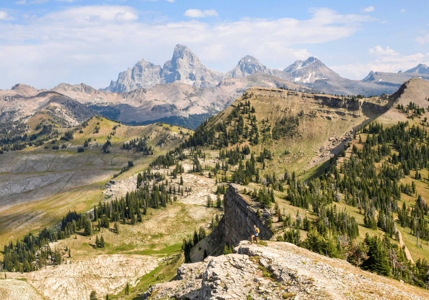 Mountain landscape with rocky ridges, green valleys, and distant peaks under a blue sky.