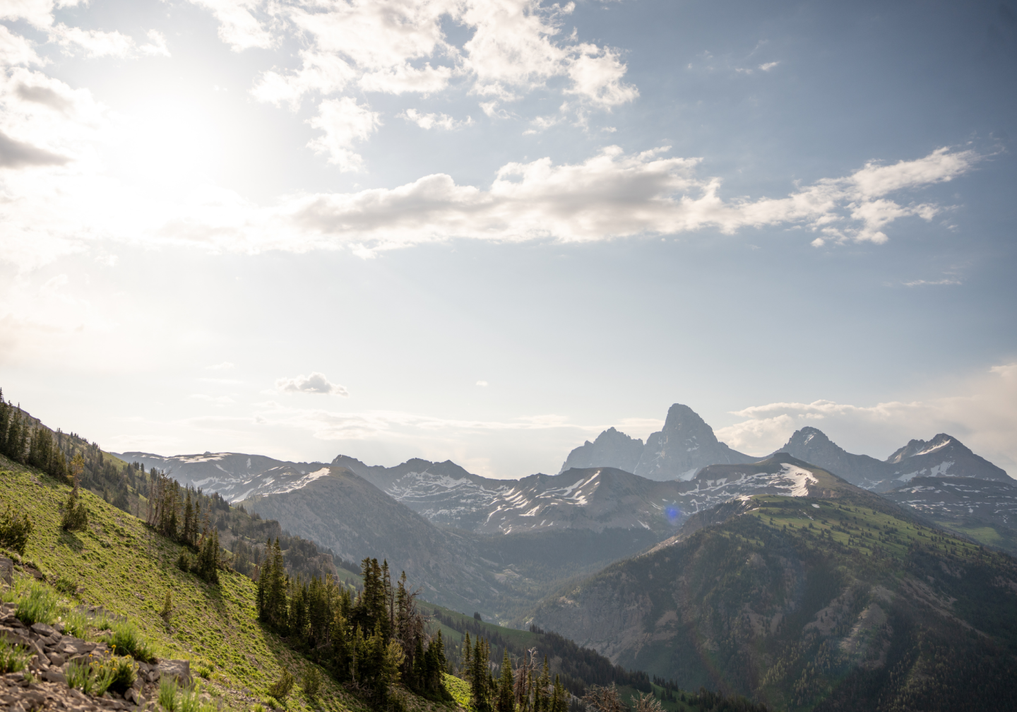 Mountain range under a bright, cloudy sky with sunlight streaming over green slopes.