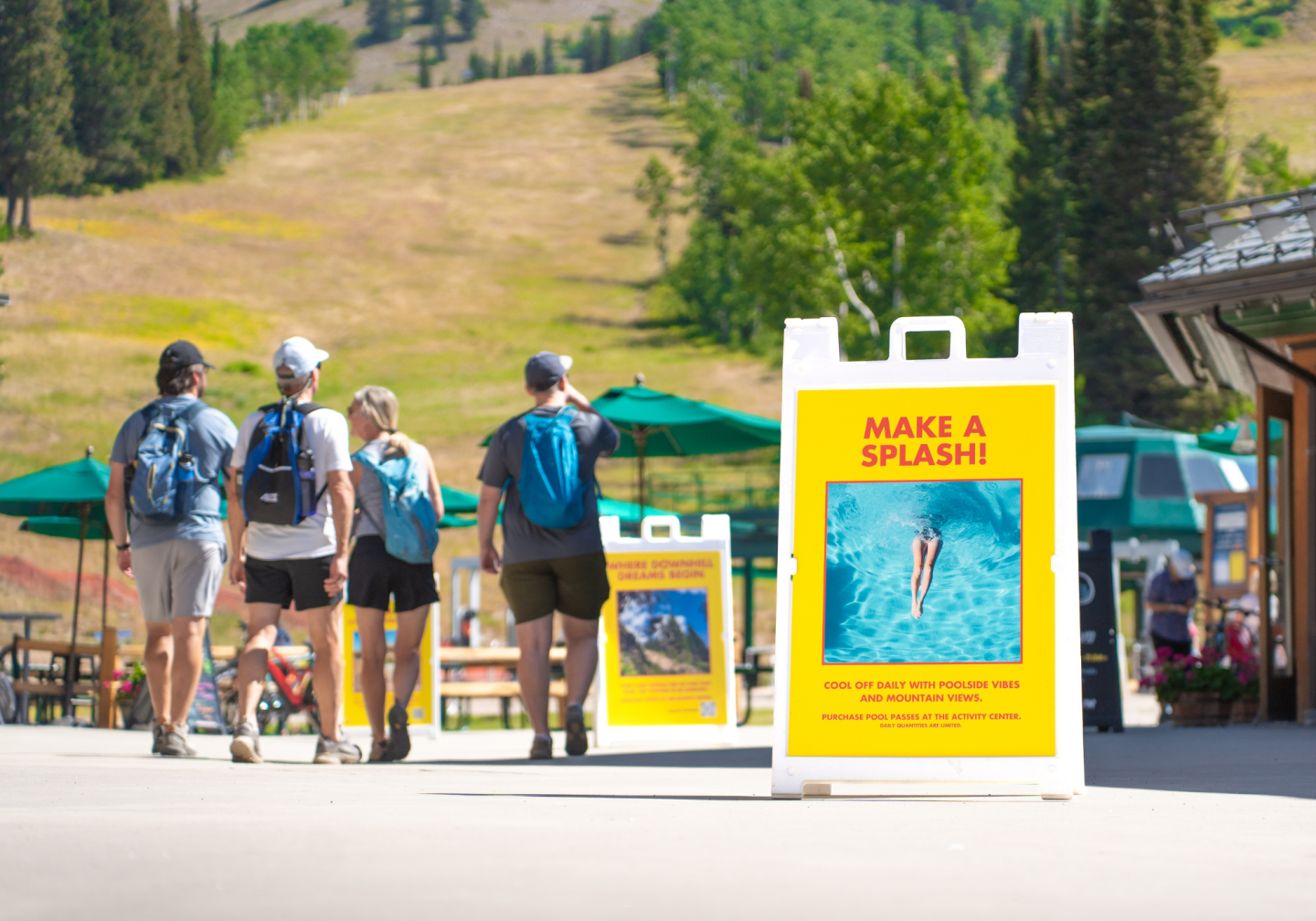 People walking on a sunny path near a park sign with hills in the background.