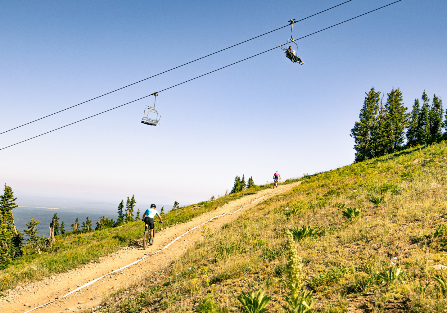 Trail on a grassy hillside with distant chairlift and trees under a clear blue sky.