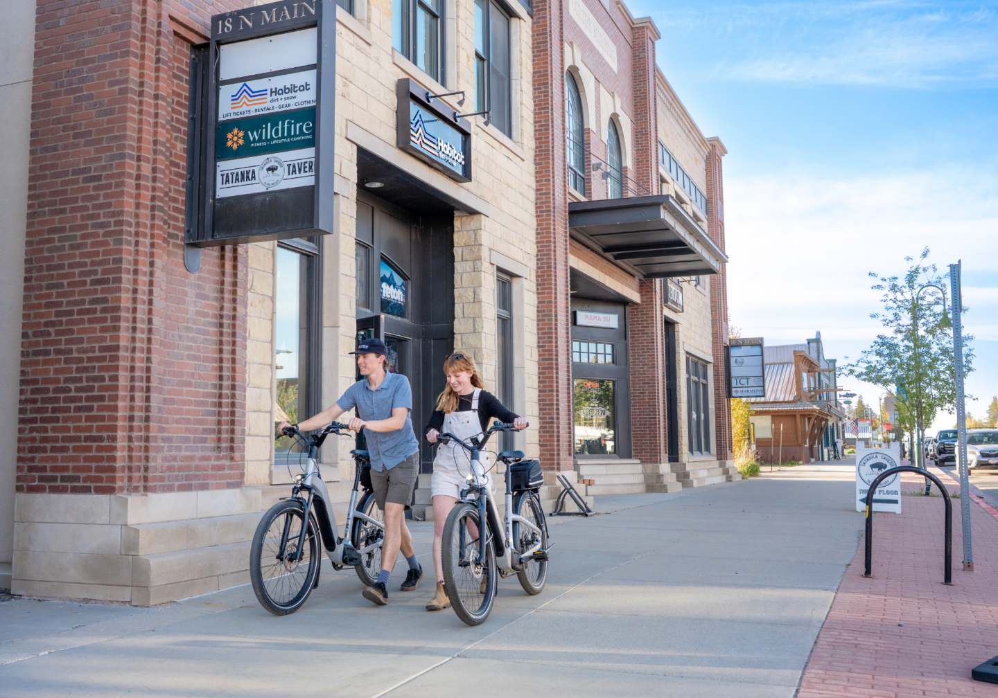 Two people walking bicycles on a sunny city sidewalk.