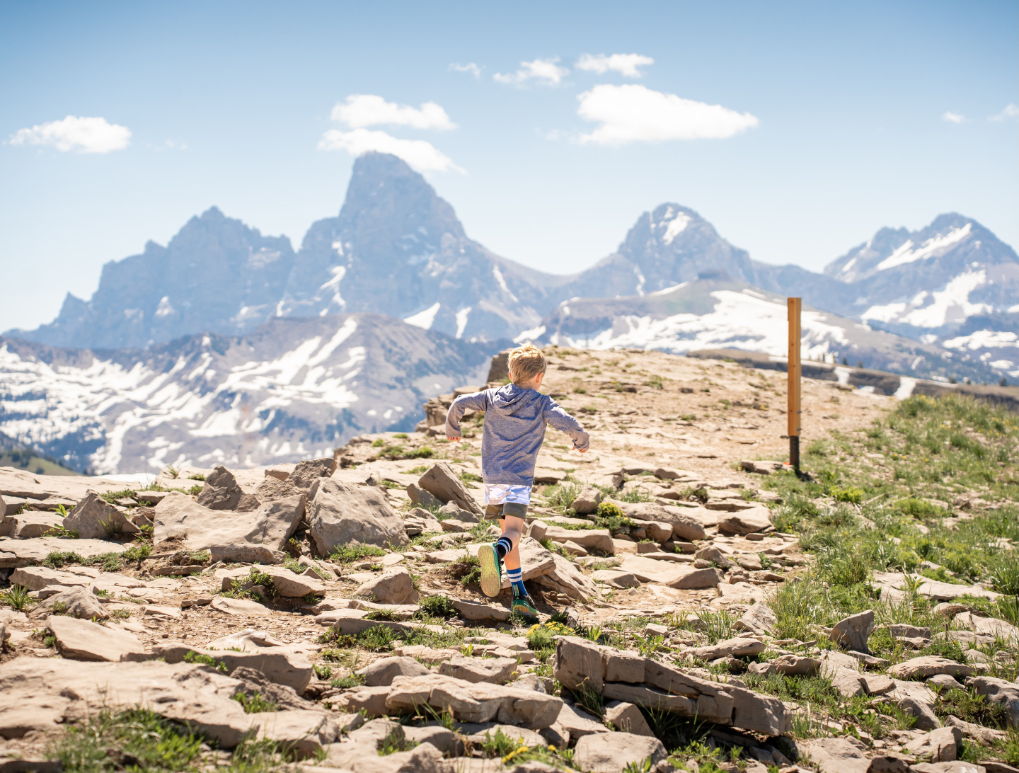 Child hiking on rocky mountain trail with distant snowy peaks.