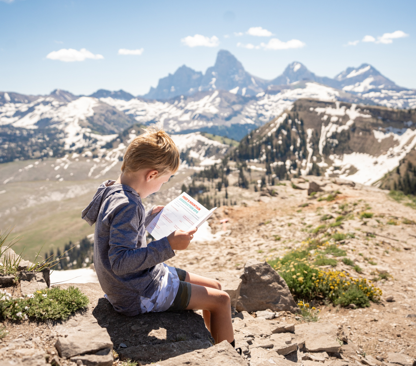 Young boy reading on a mountain ledge with snowy peaks in the background.