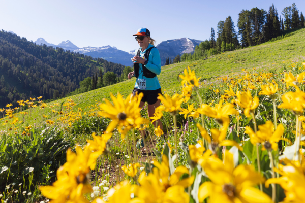 Woman hiking through a meadow of yellow flowers in sunny, mountainous landscape.