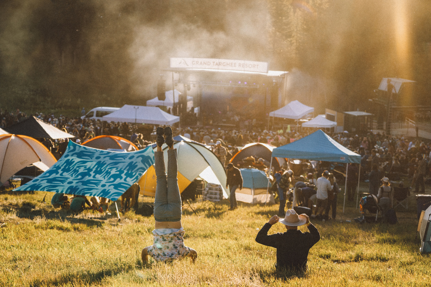 Festival scene with tents, stage, and people in a sunlit field.
