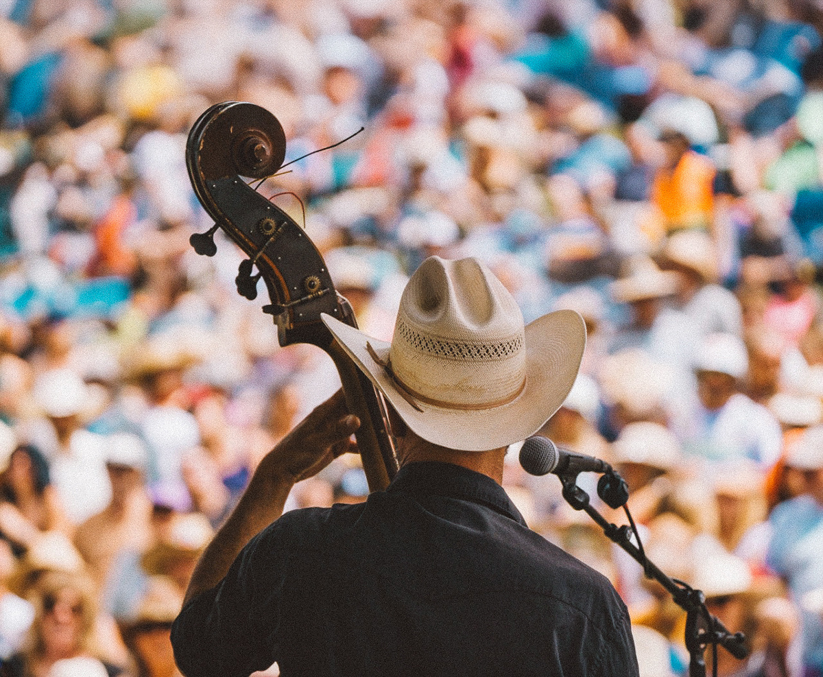 Musician in cowboy hat plays bass on stage, vibrant crowd in background.