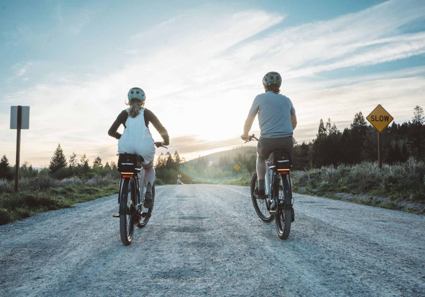 Two people cycling on a gravel path at sunset, surrounded by trees.