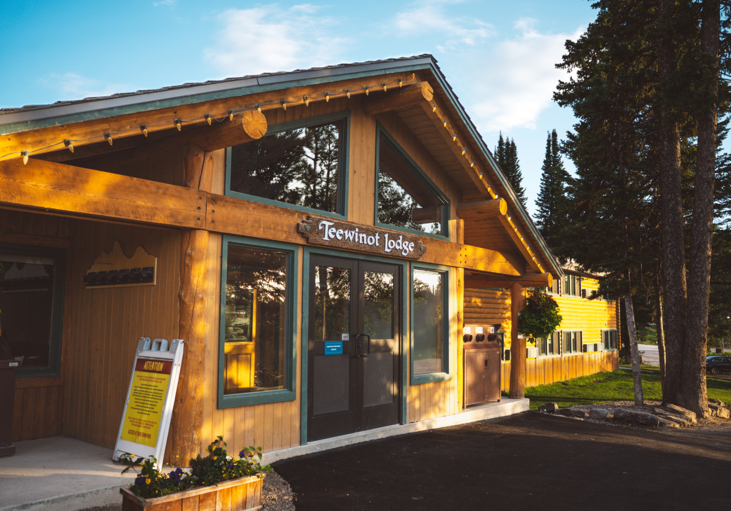 Rustic lodge entrance with large windows, surrounded by trees, under a blue sky.