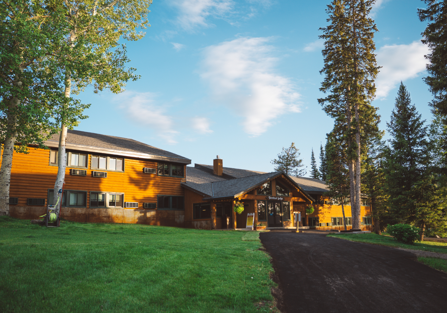 Large wooden lodge with green lawn, surrounded by tall trees under a blue sky.