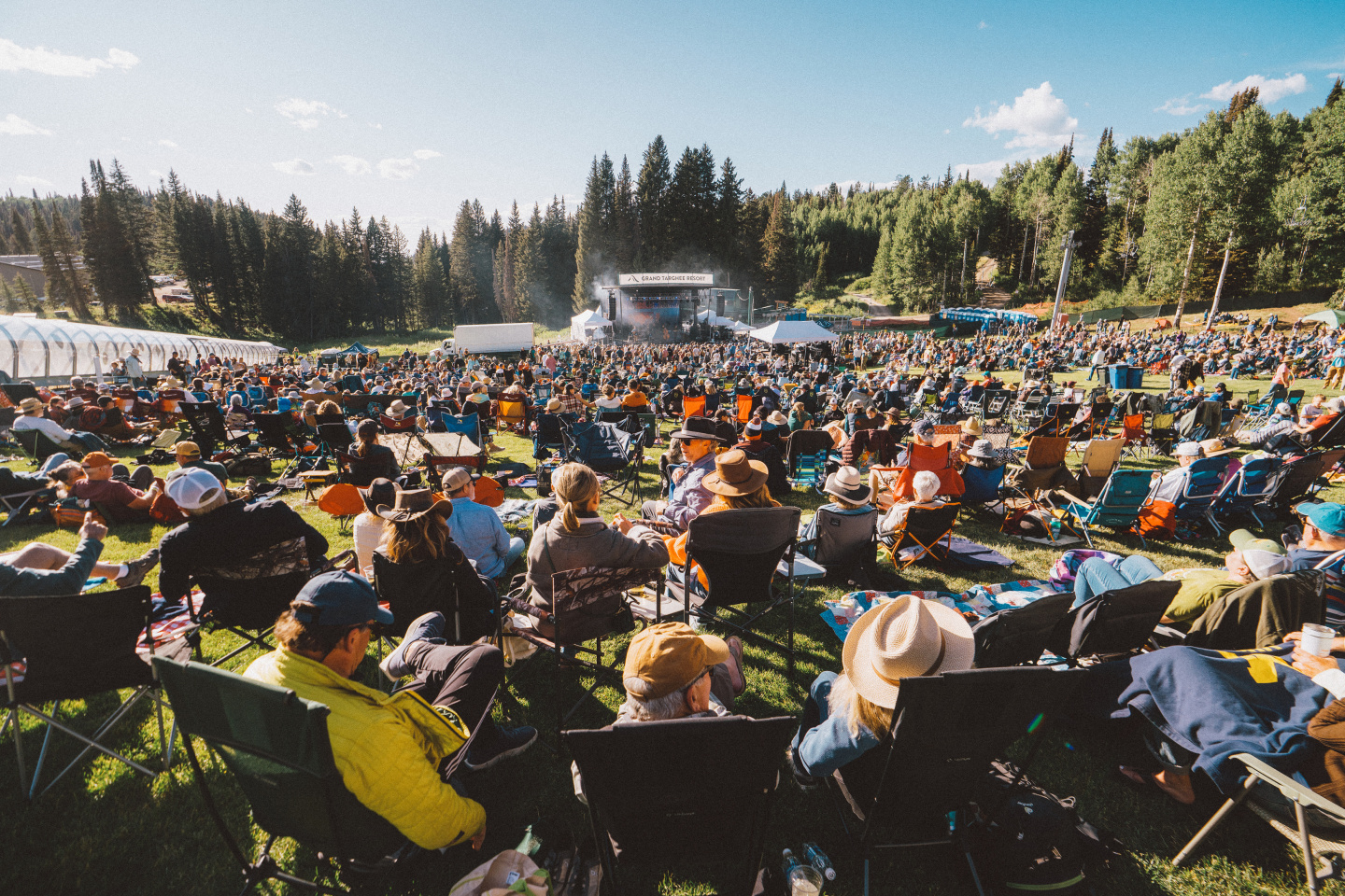 Outdoor concert with a large crowd seated on a grassy field under a clear blue sky.